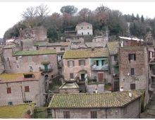 Vista panoramica di un borgo collinare con case in pietra addossate tra loro e tetti in coppi. Sullo sfondo si vede un pendio alberato con alcune abitazioni sparse e antenne sui tetti.