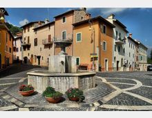 Piazza acciottolata di un borgo con una fontana in pietra al centro, circondata da vasi di fiori e panchine. Sullo sfondo si affacciano case a più piani dai colori pastello con balconi e persiane, illuminate dal sole.