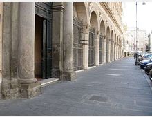 Portico urbano lungo una strada, con grandi colonne in pietra e una sequenza di archi che sostengono la facciata di un edificio storico. Sul marciapiede ampio si vedono cancellate metalliche, un ingresso con porta aperta e auto parcheggiate sul lato della carreggiata.