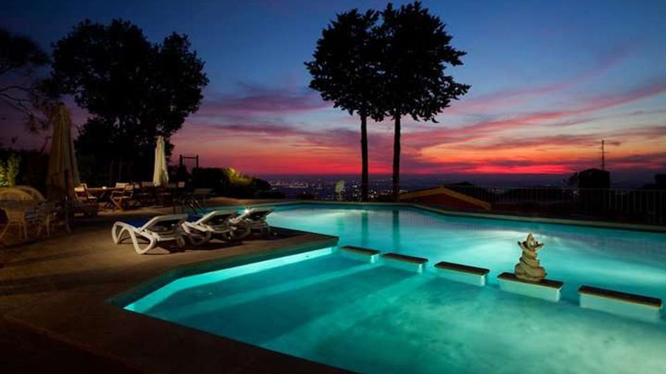 Piscina illuminata all’aperto con gradini e sedute sommerse, circondata da sdraio e ombrelloni su una terrazza. Sullo sfondo si vedono alberi in controluce e una vista panoramica verso l’orizzonte al tramonto.