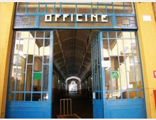 Ingresso di un edificio industriale con grandi porte vetrate blu e insegna “OFFICINE” sopra l’architrave. All’interno si vede un lungo corridoio coperto con soffitto alto ad arco e luce in fondo al passaggio.