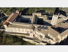 Veduta aerea di un grande complesso monastico in pietra con tetti in coppi, articolato attorno a più cortili interni e corridoi. Si notano un campanile quadrato e mura perimetrali, circondati da aree verdi e terreni agricoli.