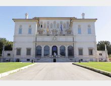 Facciata di un grande palazzo neoclassico chiaro, simmetrico, con finestre alte e tre arcate centrali al piano terra, raggiungibili da una scalinata con balaustra. Davanti si estende un viale rettilineo tra prati verdi, con bandiere sul balcone centrale e alberi ai lati sullo sfondo.