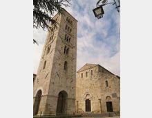 Torre campanaria medievale in pietra con alte bifore sovrapposte e grandi arcate alla base, accanto a una chiesa romanica in pietra con facciata semplice e portale ad arco. Inquadratura dal basso con cielo parzialmente nuvoloso, rami di albero e un lampione in alto a destra.