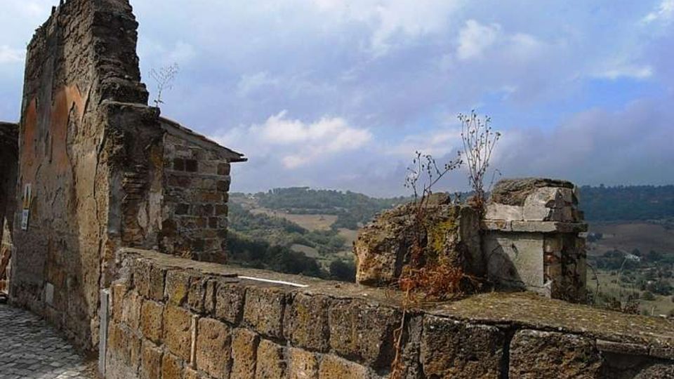 Ruderi in pietra di un edificio antico con muro basso e frammenti di muratura, affacciati su una valle collinare. Sullo sfondo si vedono campi e boschi sotto un cielo parzialmente nuvoloso.