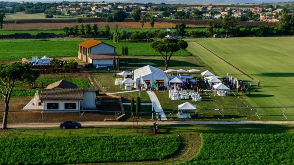 Veduta aerea di una tenuta in campagna con edifici bassi e un prato recintato allestito con grandi tensostrutture bianche, gazebo e file di sedie. Intorno si estendono campi coltivati e, sullo sfondo, un piccolo centro abitato e montagne lontane.
