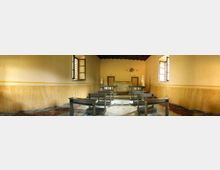Interno di una piccola aula con pareti gialle e zoccolatura in finto marmo, file di banchi in legno e pavimento in parte rovinato. In fondo si vede una pedana con cattedra e una parete decorata, con finestre laterali e soffitto scuro a travi.