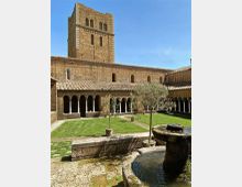 Chiostro in pietra di un complesso monastico con porticato ad archi su colonne, prato centrale e una fontana circolare in primo piano. Sullo sfondo si vede una torre campanaria rettangolare in muratura sotto un cielo sereno.