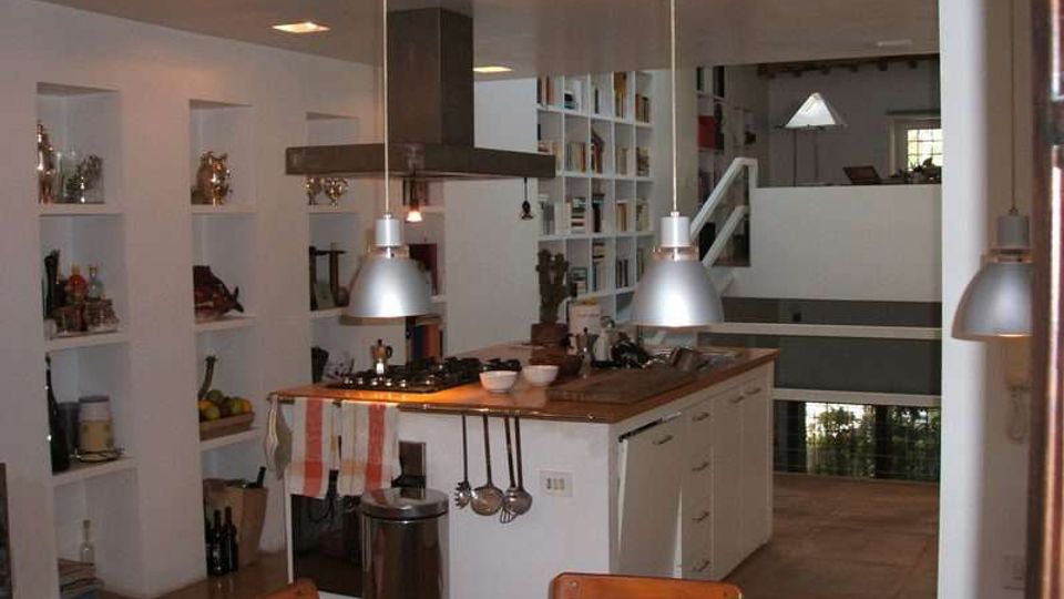 Interno di una cucina moderna a pianta aperta con isola centrale in legno, cappa a soffitto e lampade a sospensione in metallo. Pareti con nicchie e scaffalature piene di oggetti, tavolo in legno in primo piano e soppalco con ringhiera che affaccia sull’ambiente.