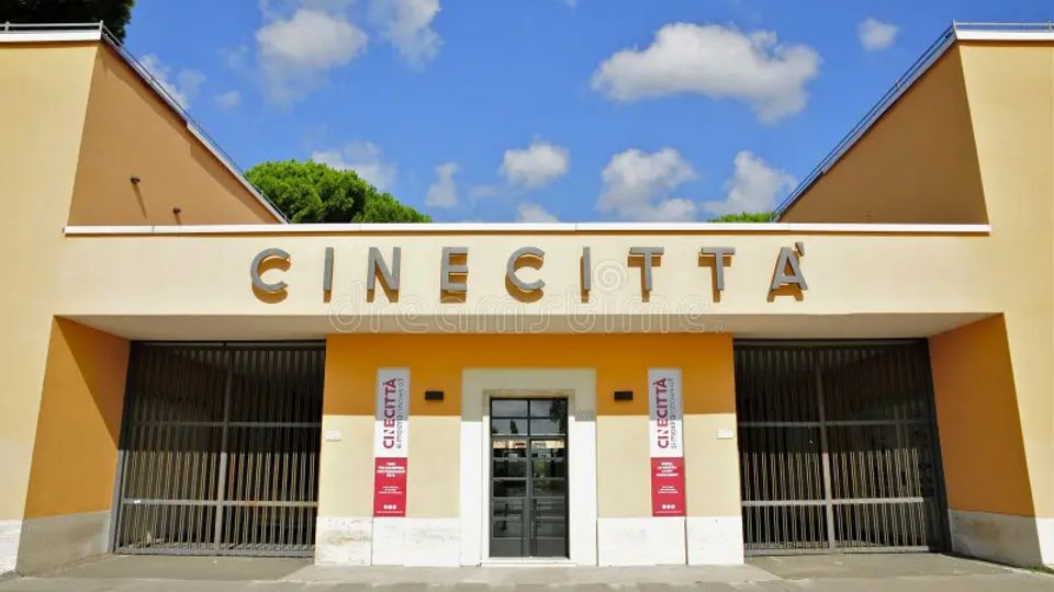 Ingresso degli Studi di Cinecittà, con facciata simmetrica color crema e ocra e grande scritta “CINECITTÀ” in alto. Al centro c’è un portone scuro affiancato da due banner verticali, con ampie cancellate metalliche ai lati e cielo azzurro con nuvole sullo sfondo.