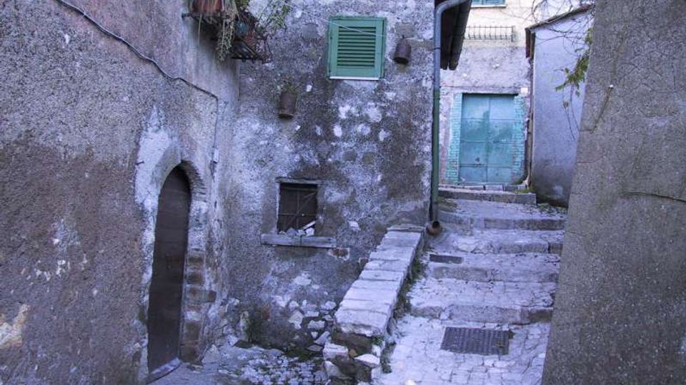 Vicolo stretto in pietra con gradini che salgono tra case antiche intonacate e muri invecchiati. Si vedono una porta ad arco, piccole finestre con persiane verdi e una grande porta verde in fondo al passaggio.