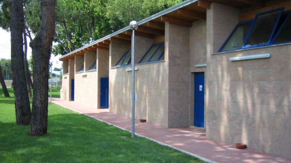 Edificio basso e allungato in muratura chiara, con tetto a falda sporgente e finestre orizzontali inclinate sopra una serie di porte blu. Davanti corre un camminamento in mattoni affiancato da prato e pini, con un lampione al centro e alberi sullo sfondo.