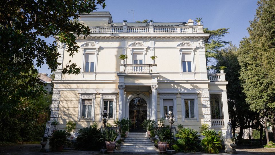 Facciata di una villa storica a due piani intonacata chiara, con ingresso ad arco e colonne, balcone centrale e ringhiere decorative sul tetto. Davanti, una scalinata con vasi e piante ornamentali conduce all’entrata, circondata da alberi e giardino.