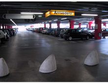 Parcheggio multipiano coperto con file di auto parcheggiate ai lati, colonne rosse e soffitto basso con illuminazione a neon. In alto sono visibili cartelli gialli di segnaletica, tra cui “Arrivederci” e “Uscita”.