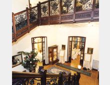 Interno di un elegante atrio su due livelli, con balconata in legno e ringhiere in ferro battuto decorato. Al piano terra si vedono pavimento in parquet con tappeti, vetrina espositiva, quadri alle pareti e due porte ad arco che conducono a sale apparecchiate per banchetti.