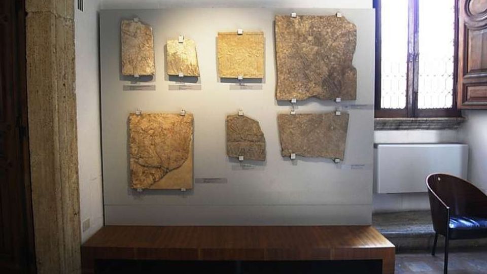Pannello espositivo in una sala museale con grandi frammenti di pietra o lastre antiche fissati alla parete sopra una pedana in legno. Accanto si vede una finestra alta con inferriate e una parete in pietra, con pavimento in piastrelle.