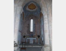 Interno di una piccola cappella in pietra con abside e altare centrale, colonne laterali e affreschi sulle pareti e sulla volta. Sul fondo una stretta finestra ad arco illumina l’altare con candelabri e vasi di piante.