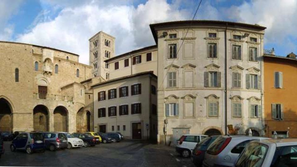 Piazza urbana con parcheggio di auto, circondata da edifici storici: una chiesa in pietra con archi e portali a sinistra e palazzi residenziali a più piani con facciate intonacate e finestre regolari a destra. Sullo sfondo si vede un campanile in pietra sotto un cielo parzialmente nuvoloso.