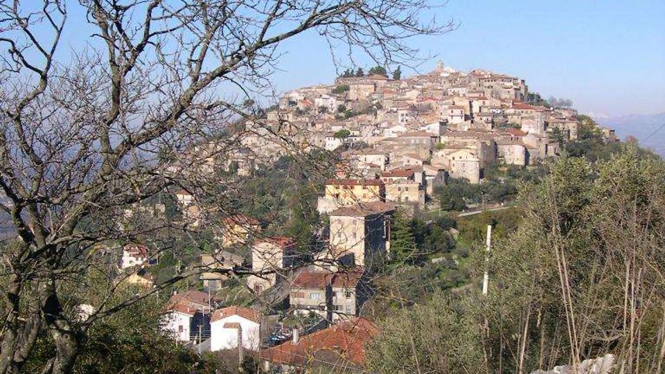 Veduta panoramica di un borgo collinare con case in pietra e tetti in coppi, disposto a gradoni sul pendio. In primo piano ci sono rocce, erba e rami di alberi spogli, con vegetazione e cielo sereno sullo sfondo.