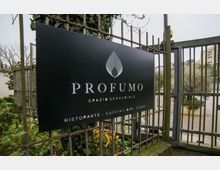 Insegna nera con il logo e la scritta “PROFUMO – Spazio sensoriale” fissata a una recinzione metallica vicino a un cancello d’ingresso. Sullo sfondo si vedono vegetazione, un vialetto e edifici lontani.