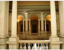Interno di un cortile coperto con colonne in pietra e logge ad archi su due livelli, con balaustre e portali in legno sullo sfondo. Illuminazione calda sulle pareti e dettagli classici lungo i cornicioni.