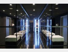 Bagno moderno con pareti specchiate e illuminazione a LED verticali lungo la parete centrale, che riflettono su pavimento scuro lucido e soffitto con faretti incassati. Ai lati ci sono lavabi bianchi squadrati con rubinetti cromati e porte su entrambe le estremità del locale.