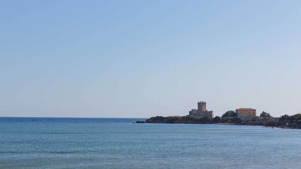 Veduta costiera con mare calmo in primo piano e, in lontananza, un piccolo promontorio con una torre in pietra e alcuni edifici. Cielo sereno sopra l’orizzonte marino.