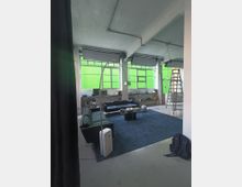 Interno di un ampio loft con soffitto in cemento a vista e grandi finestre a riquadri schermate da pannelli verdi. Area soggiorno con divani, tavolino basso su tappeto blu, lampada ad arco e una scala a pioli vicino alle finestre.