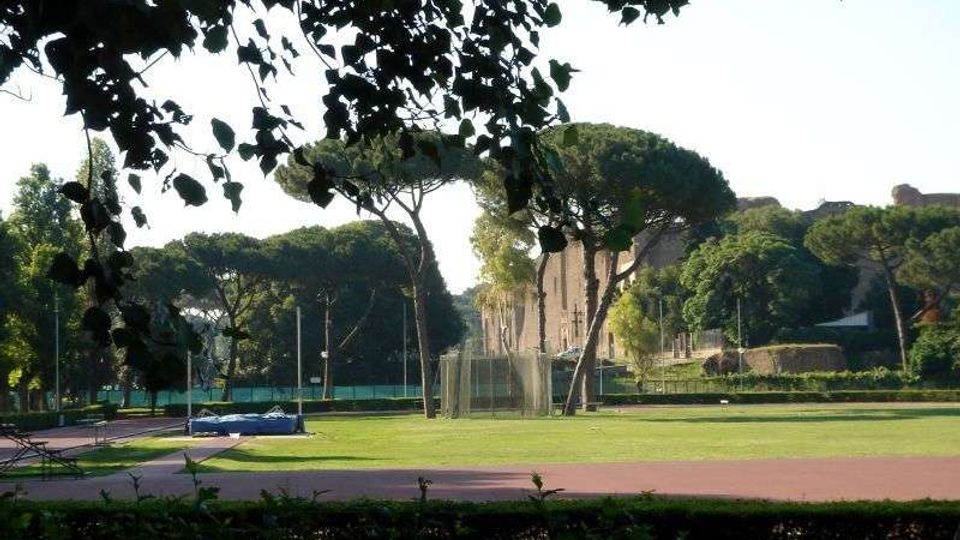 Campo sportivo all’aperto con pista rossa perimetrale e ampia area erbosa centrale, circondato da siepi e alti pini a ombrello. Sullo sfondo si vedono recinzioni, reti e una struttura in muratura parzialmente visibile tra gli alberi.