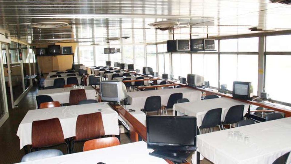 Ampia sala interna con file di tavoli rettangolari coperti da tovaglie bianche e sedie, con numerosi monitor e televisori distribuiti sui tavoli e lungo le pareti. Grandi finestre laterali illuminano l’ambiente, con soffitto metallico e ventilatori circolari.
