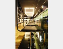Interno di un bar moderno con fila di sgabelli alti in metallo e sedute scure lungo un bancone con parete in vetro. Illuminazione a soffitto con faretti e grande lampada rettangolare sospesa, pavimento in legno e superfici lucide.