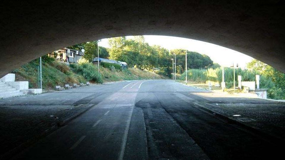 Strada asfaltata vista dall’interno di un sottopasso ad arco, con corsia ciclabile centrale e carreggiata che prosegue in leggera curva. Ai lati si vedono scarpate erbose, alberi e alcuni lampioni lungo il margine della strada.