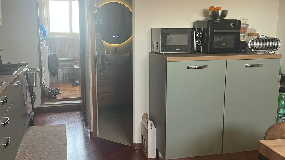 Interno di una cucina moderna con pavimento in legno scuro, mobili verde salvia e piano in legno con microonde e piccoli elettrodomestici. A sinistra c’è una fila di basi cucina e, in fondo, una porta conduce a un bagno con specchio rotondo illuminato e a una zona luminosa con finestra.