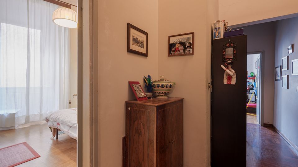 Corridoio interno di un appartamento con pavimento in legno, un mobile basso in legno con oggetti decorativi e quadri alle pareti, affiancato da un armadio scuro. A sinistra si apre una stanza luminosa con grandi tende bianche e lampada a sospensione, mentre a destra il corridoio prosegue verso altre stanze.