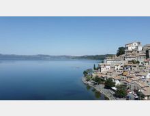 Veduta panoramica di un grande lago calmo con colline basse all’orizzonte. Sulla riva a destra si trova un borgo compatto di case in pietra con tetti in coppi, disposto in salita fino a un edificio più grande in cima, con una strada e alberi lungo il lungolago.