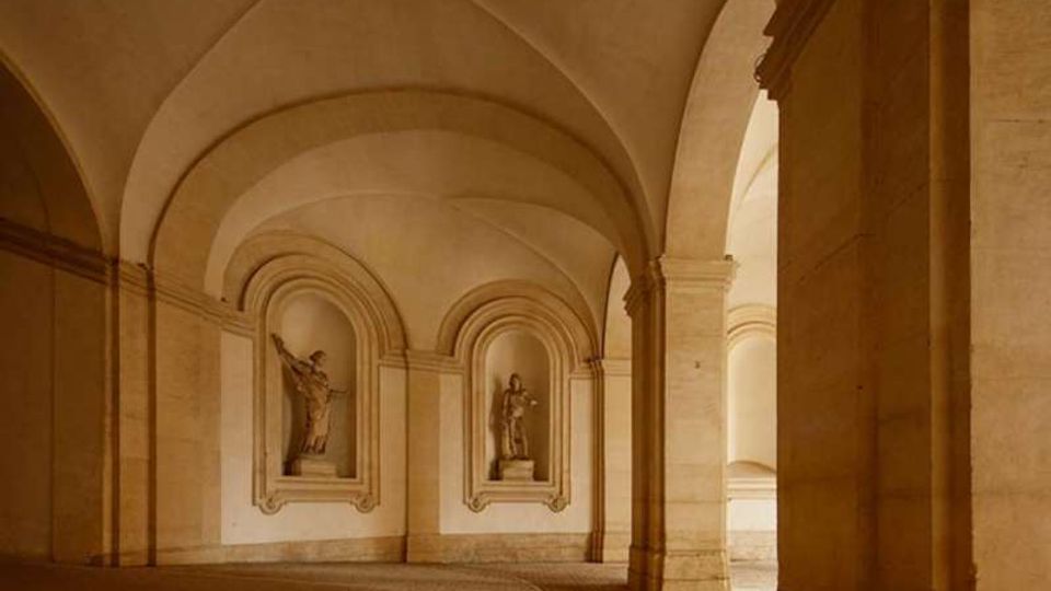 Interno di un porticato in pietra con volte a botte e archi, sostenuto da pilastri massicci. Sulla parete di fondo si vedono due nicchie ad arco con statue e pavimento in pietra.