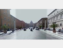 Ampio viale urbano lastricato con traffico e auto parcheggiate ai lati, fiancheggiato da palazzi storici in muratura e lampioni. In fondo alla prospettiva si vede la cupola della Basilica di San Pietro.