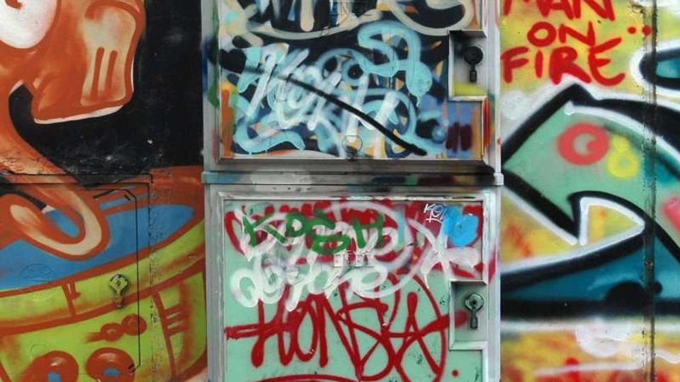 Armadio tecnico metallico da strada ricoperto di graffiti e tag, posizionato davanti a un muro con murales multicolori e scritte spray. In primo piano si vede il marciapiede in cemento e mattonelle con piccoli detriti.