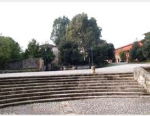 Ampia scalinata in pietra che conduce a una piazza lastricata, circondata da alberi e lampioni. Sullo sfondo si vedono edifici in muratura, tra cui una struttura in mattoni rossi con archi e un ingresso.