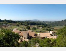 Veduta panoramica di un complesso di edifici rurali con tetti in tegole color terracotta, circondato da alberi e strade sterrate. Sullo sfondo si estendono colline coltivate e campi verdi fino a montagne lontane sotto un cielo sereno.