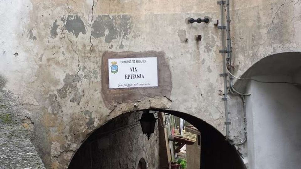 Stretto vicolo in pietra che scende sotto un arco ricavato tra edifici antichi con intonaco consumato e crepe. Sulla facciata si vedono persiane verdi e un cartello stradale “Via Epifania”, con cavi esterni e una piccola apertura circolare nel muro.
