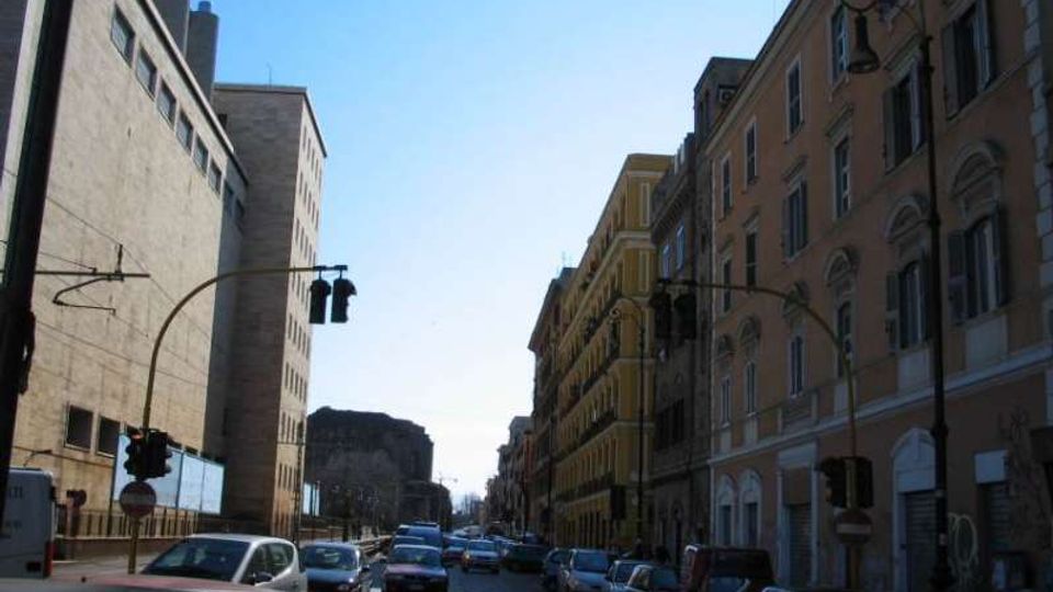Strada urbana rettilinea con molte auto in sosta e in transito, fiancheggiata da edifici alti: a destra palazzi storici con finestre ad arco, a sinistra un grande edificio moderno. Sono visibili semafori e lampioni lungo la carreggiata sotto un cielo sereno.