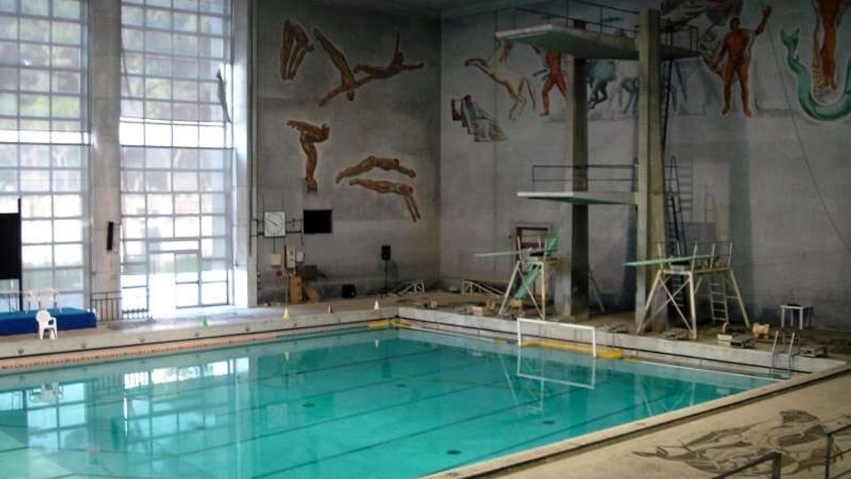 Piscina coperta con grande vasca rettangolare e piattaforme per tuffi su una torre, circondata da bordo in cemento e corrimano. Alte finestre a griglia illuminano l’ambiente e sulle pareti sono dipinti murales con figure di tuffatori e nuotatori.