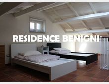 Camera da letto mansardata con soffitto in legno a travi bianche, pavimento in cotto e due letti bassi con comodino; una finestra con veneziane illumina l’ambiente vicino a una ringhiera interna.