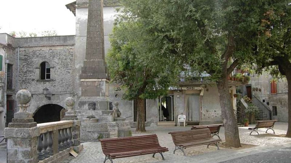 Piccola piazza lastricata con panchine in legno e alberi ombrosi, dominata da un alto obelisco monumentale su basamento in pietra accanto a una balaustra. Sullo sfondo si vedono edifici in muratura e intonaco con arcate e ingressi al piano terra.