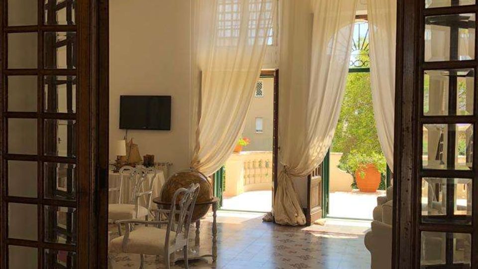 Interno di un salone luminoso visto attraverso una grande porta in legno e vetro, con pavimento in piastrelle decorate, tavolo con sedie e arredi classici. Alte tende chiare incorniciano porte-finestre aperte su un balcone con balaustra e grandi vasi di piante.