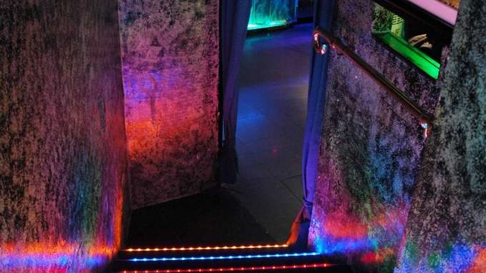 Scala interna in un corridoio stretto con pareti scure, gradini illuminati da strisce LED multicolore e un arco che conduce a un’area con luci blu. Sulla parete destra è incassato un acquario retroilluminato con cornice luminosa.