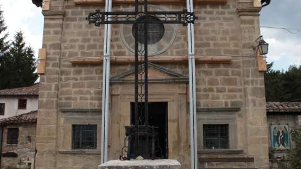 Facciata frontale di una piccola chiesa in pietra con tetto a capanna e ingresso centrale, con finestre laterali e un oculo sopra il portale. Davanti all’edificio si trova una grande croce in ferro su un basamento in muratura, in un cortile con pavimentazione in pietra e prato sotto un cielo nuvoloso.