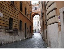 Stretto vicolo urbano in pietra con pavimentazione a sampietrini, fiancheggiato da alti edifici storici in muratura. In fondo si apre un arco che conduce verso una strada più ampia con facciate monumentali visibili oltre il passaggio.