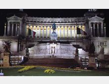 Facciata monumentale illuminata di un grande edificio neoclassico con lunga fila di colonne e due avancorpi laterali con timpani. Al centro si vede una statua equestre su alto basamento, con scalinata e giardino in primo piano e bandiere italiane ai lati.